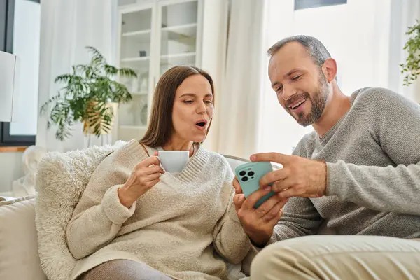 Чоловік показує смартфон здивованій дружині з чашкою кави на дивані у вітальні, бездітна пара — стокове фото