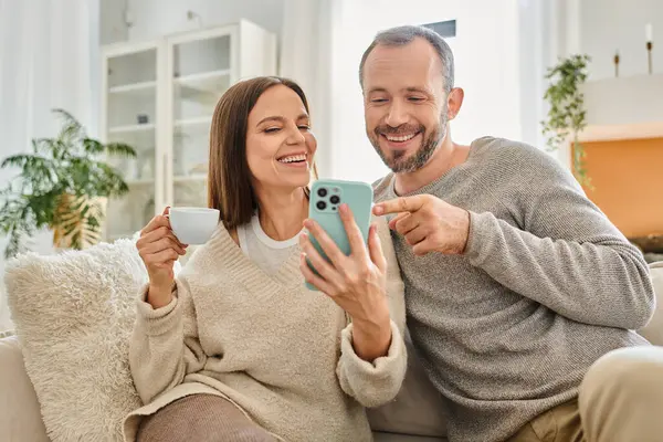 Hombre alegre apuntando al teléfono inteligente cerca de la esposa con taza de café en el sofá en casa, estilo de vida libre de niños - foto de stock