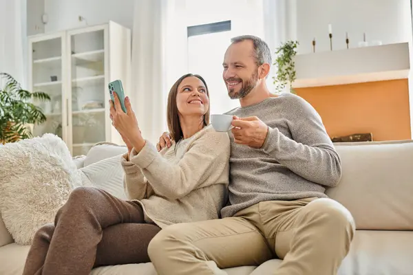 Femme joyeuse montrant smartphone au mari avec tasse de café sur le canapé dans le salon, vie sans enfant — Photo de stock