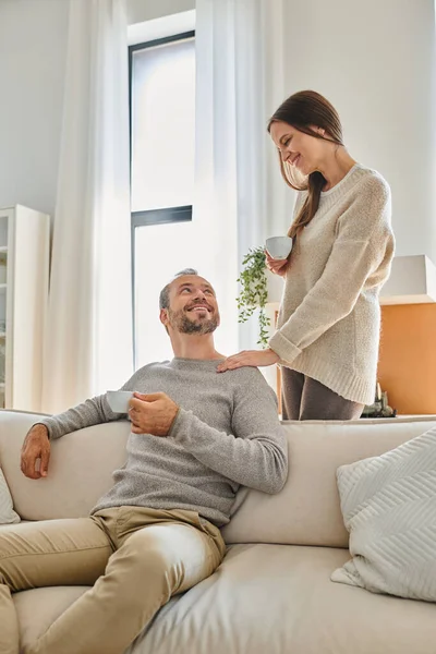 Child-free couple with coffee cups smiling at each other in cozy living room at home, happy morning — Stock Photo