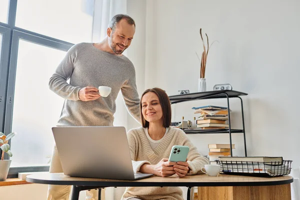 Sorrindo homem com copo de café apoiando esposa trabalhando no laptop em casa, unidade de casal livre de crianças — Fotografia de Stock
