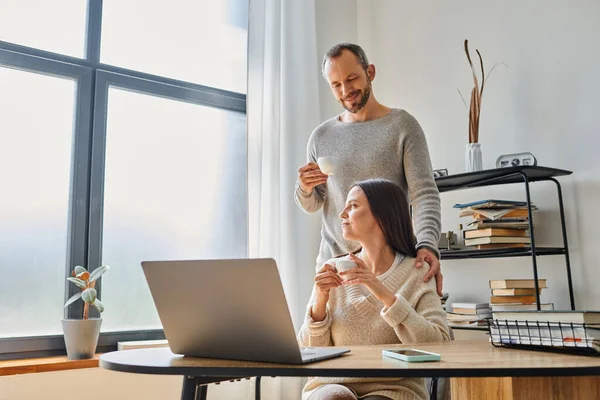 Marido cariñoso con taza de café esposa de apoyo que trabaja en el ordenador portátil en casa, la unidad de la pareja libre de niños — Stock Photo