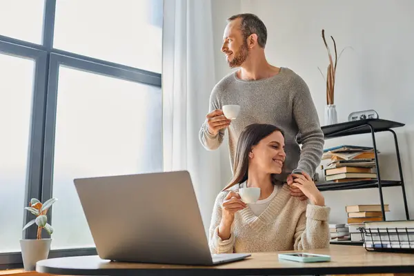 Улыбающийся мужчина с кофейной чашкой, поддерживающей жену, работающую на ноутбуке дома, единство бездетной пары — стоковое фото