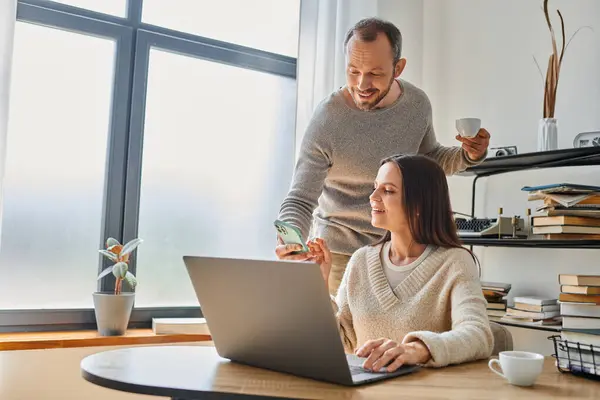 Sorrindo homem com xícara de café mostrando smartphone para esposa trabalhando no laptop em casa, casal sem filhos — Fotografia de Stock