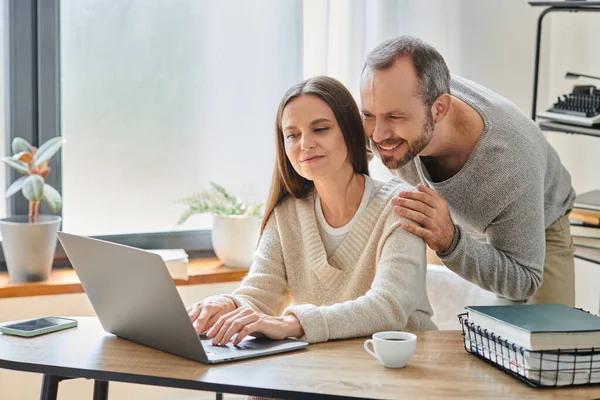 Счастливый мужчина обнимает плечи улыбающейся жены, работающей на ноутбуке дома, без детей образ жизни — стоковое фото