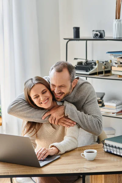 Uomo amorevole che abbraccia la moglie sorridente che lavora sul computer portatile a casa, la cura e il sostegno della coppia senza figli — Foto stock
