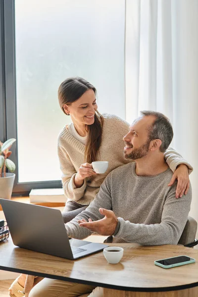 Happy woman with coffee cup supporting husband working on laptop in home office, child-free couple — Stock Photo