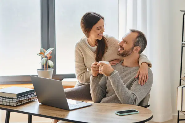 Счастливый мужчина с чашкой кофе, сидящий рядом с ноутбуком и заботливая жена в домашнем офисе, образ жизни без детей — стоковое фото
