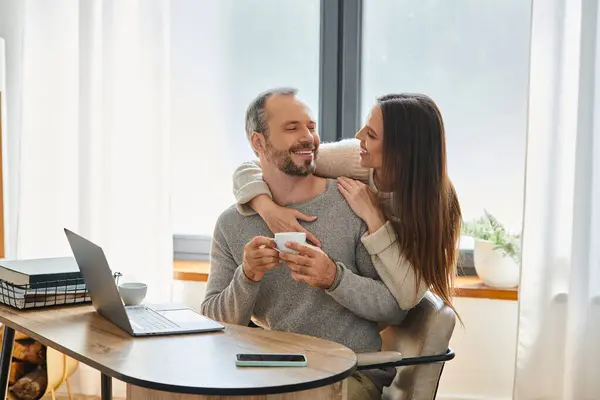 Hombre alegre con taza de café sentado cerca de la computadora portátil y la esposa cariñosa en la oficina en casa, pareja libre de niños - foto de stock
