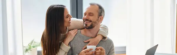 Alegre mujer abrazando sonriente marido sosteniendo taza de café cerca de la computadora portátil, pancarta horizontal - foto de stock