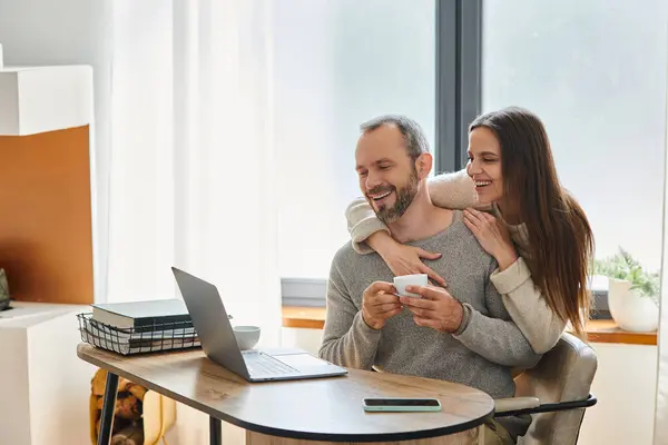 Mujer alegre abrazando marido sonriente sentado con taza de café cerca de la computadora portátil, estilo de vida libre de niños - foto de stock