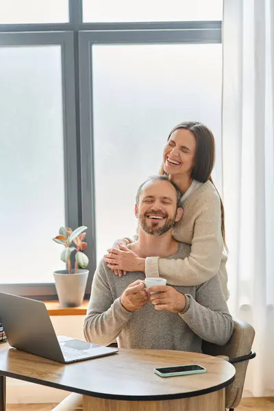 Взволнованная жена обнимает улыбающегося мужа, сидящего с чашкой кофе возле ноутбука, образ жизни без детей — стоковое фото