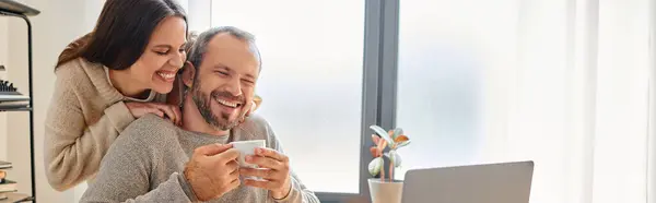 Mujer alegre abrazando marido feliz sosteniendo taza de café cerca de la computadora portátil, estilo de vida libre de niños, bandera - foto de stock