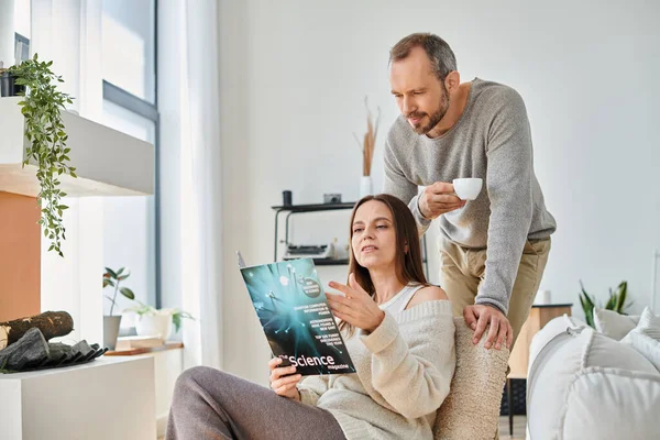 Mujer leyendo revista de ciencias cerca de marido con taza de café en el sofá en casa, pareja libre de niños - foto de stock