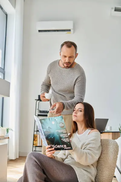 Hombre con taza de café apuntando a la revista científica cerca de alambre de lectura en el sofá en acogedora sala de estar - foto de stock