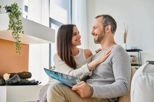 Smiling woman embracing husband reading science magazine on couch at home, child-free life — Stock Photo