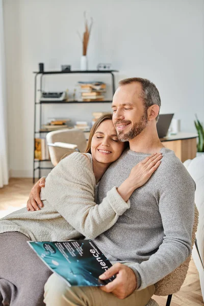 Delighted woman hugging husband sitting with science magazine on couch at home, child-free life — Stock Photo