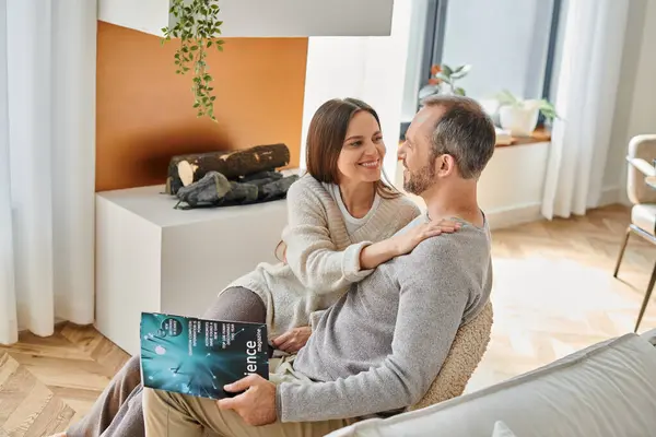 Mujer alegre abrazando marido sentado con la revista de ciencia en el sofá en casa, pareja libre de niños - foto de stock