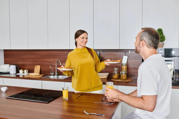 Mujer feliz sosteniendo delicioso desayuno cerca del marido con jugo de naranja en la cocina, vida libre de niños - foto de stock