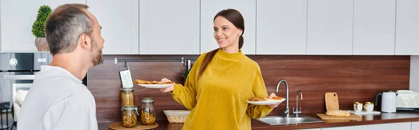 Sonriente mujer sosteniendo delicioso desayuno cerca complacido marido en cocina, vida libre de niños, pancarta - foto de stock