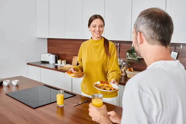 Joyeuse femme tenant un délicieux petit déjeuner près du mari avec du jus d'orange dans la cuisine, couple sans enfant — Photo de stock
