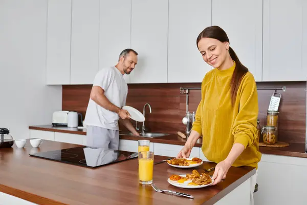 Lächelnde Frau serviert leckeres Frühstück, während Ehemann Geschirr in Küche spült, Pflege und Harmonie — Stockfoto