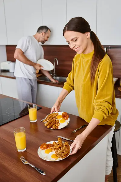 Donna gioiosa che serve gustosa colazione vicino al marito lavare i piatti in cucina, cura e armonia — Foto stock