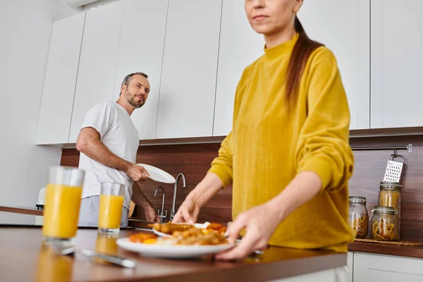 Жінка, що подає сніданок біля чоловіка миє посуд на кухні, гармонія бездітної пари — стокове фото