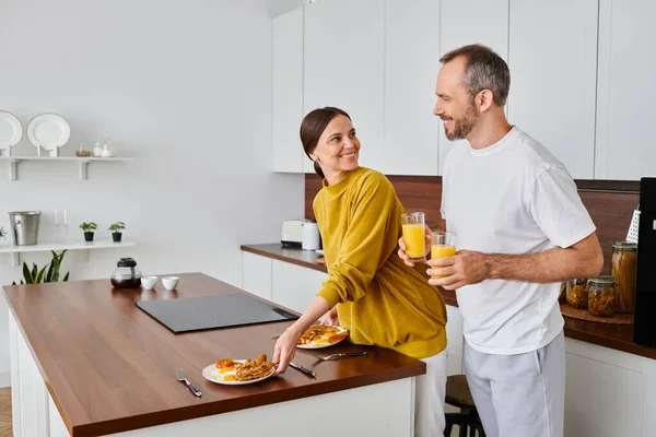 Fröhliche Frau serviert Frühstück in der Nähe ihres Mannes mit Orangensaft in der Küche, kinderfreier Lebensstil — Stockfoto