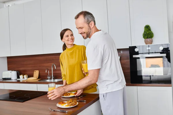 Freudige Frau serviert Frühstück in der Nähe Ehemann mit Orangensaft in der Küche, kinderfreier Lebensstil — Stockfoto