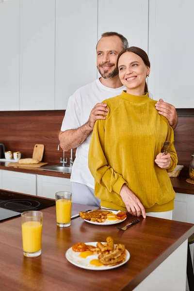 Усміхнений чоловік обіймає плечі дружини біля смачного сніданку на кухні, безкоштовна пара — стокове фото