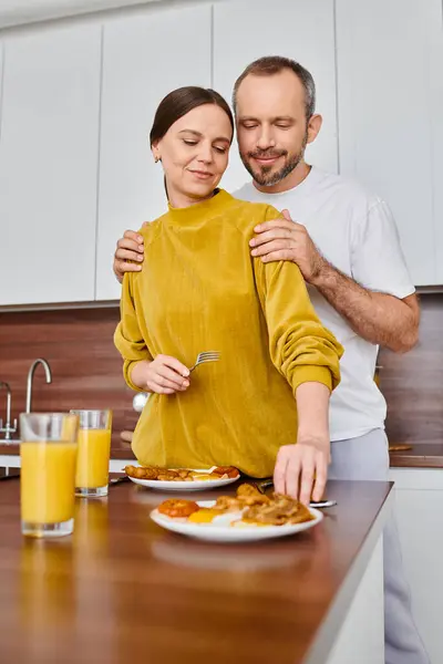 Щасливий чоловік обіймає плечі дружини біля смачного сніданку на кухні, безкоштовна пара — стокове фото