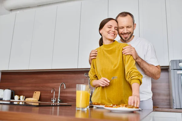 Joyful man with closed eyes hugging shoulders of wife near breakfast in kitchen, child-free couple — Stock Photo