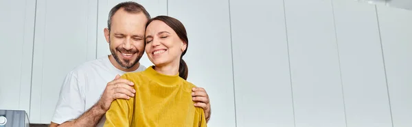 Marido cariñoso con los ojos cerrados abrazando hombros de esposa feliz en la cocina, bandera horizontal - foto de stock