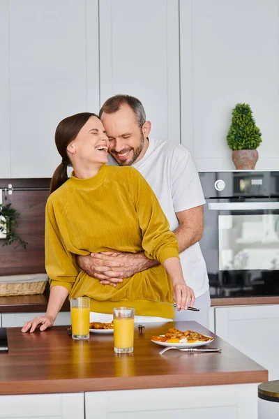 Happy man embracing laughing wife near delicious breakfast in kitchen, morning of child-free couple — Stock Photo