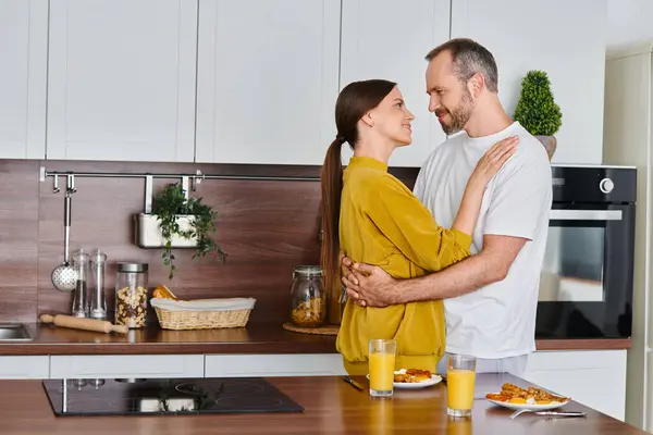 Alegre pareja sin niños abrazando cerca de delicioso desayuno en la cocina moderna, el amor y el cuidado - foto de stock