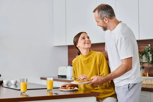 Husband serving delicious breakfast near excited wife in kitchen, morning of child-free couple — Stock Photo