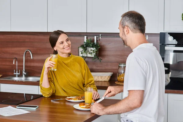 Mujer complacida sosteniendo jugo de naranja durante el desayuno con el marido, mañana de pareja libre de niños - foto de stock
