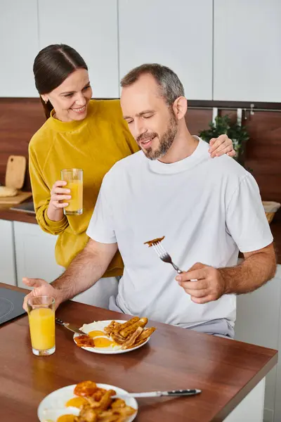 Happy wife holding orange juice near husband enjoying tasty breakfast in kitchen, love and care — Stock Photo