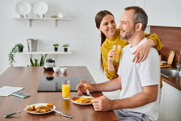 Cheerful wife holding orange juice near husband enjoying breakfast in kitchen, child-free couple — Stock Photo