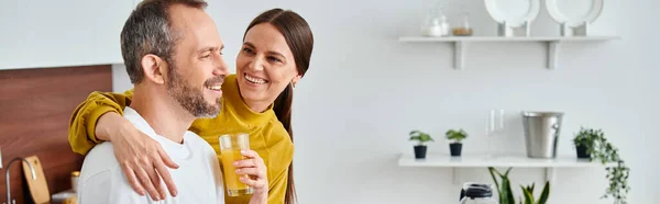 Заботливая жена держит свежий домашний апельсиновый сок рядом с восторженным мужем на кухне, баннер — стоковое фото
