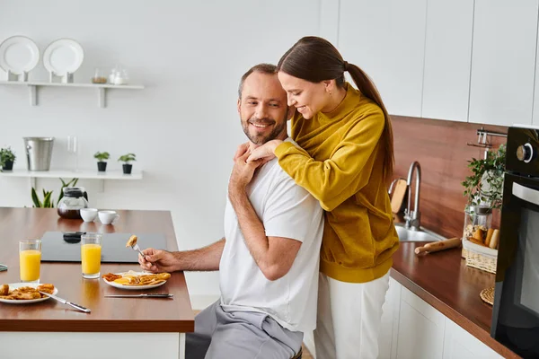 Seitenansicht eines lächelnden Mannes, der leckeres Frühstück in der Nähe seiner fürsorglichen Frau in der Küche genießt, kinderfreies Paar — Stockfoto