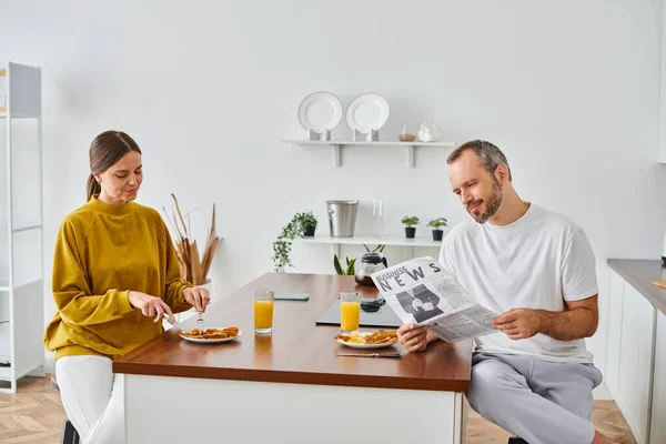 Hombre positivo leyendo noticias de negocios cerca de la esposa disfrutando de sabroso desayuno en la cocina, pancarta horizontal - foto de stock