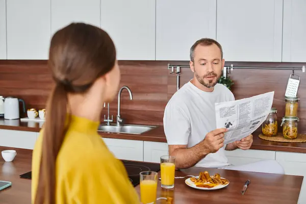 Sorprendido hombre leyendo el periódico cerca de la esposa disfrutando de sabroso desayuno en la cocina, pareja libre de niños - foto de stock