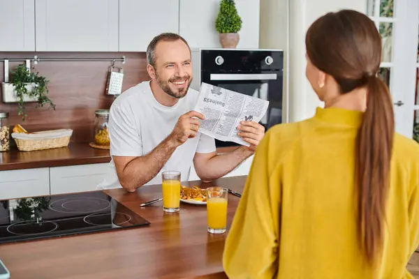 Hombre alegre mostrando periódico a la esposa durante el desayuno en la cocina moderna, estilo de vida libre de niños - foto de stock