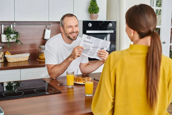 Hombre alegre mostrando periódico a la esposa durante el desayuno en la cocina moderna, estilo de vida libre de niños - foto de stock