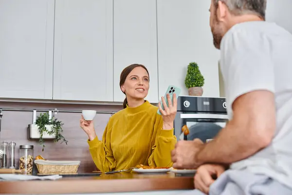 Excited woman with smartphone and coffee cup talking to husband during breakfast in kitchen — Stock Photo
