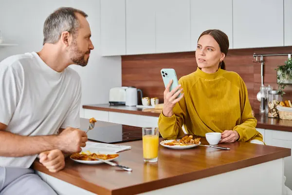 Усвідомлена жінка тримає смартфон біля чоловіка під час сніданку на кухні, безкоштовна пара — стокове фото