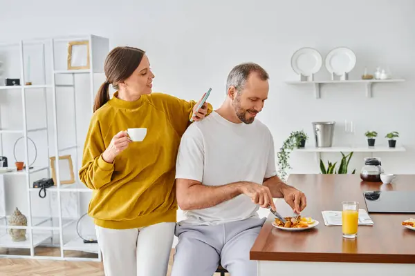 Hombre deleitado disfrutando de delicioso desayuno cerca de la esposa con taza de café y teléfono inteligente en la cocina - foto de stock