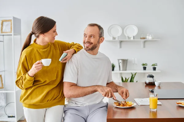 Hombre alegre disfrutando de sabroso desayuno cerca de la esposa con taza de café y teléfono inteligente en la cocina moderna - foto de stock